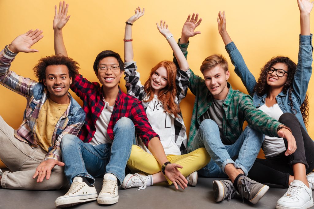 Multiethnic group of cheerful young people sitting with raised hands and having fun over yellow background